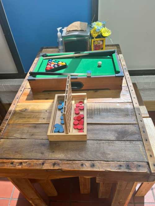 A wooden table with a small pool table, game pieces, and a vase of flowers in a bright room.