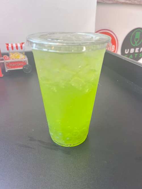 A clear cup filled with a bright green drink and ice, featuring tapioca pearls at the bottom, on a black tray.