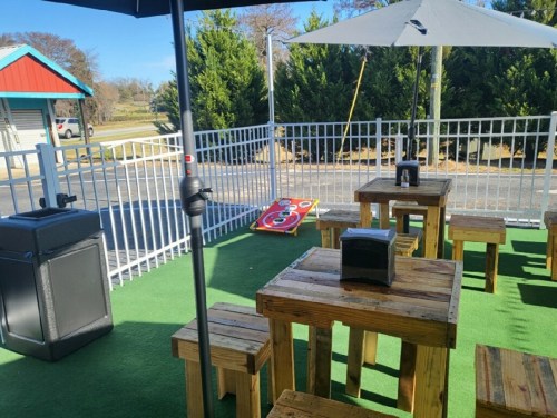 Outdoor seating area with wooden tables, an umbrella, and a cornhole game on a green turf surface.