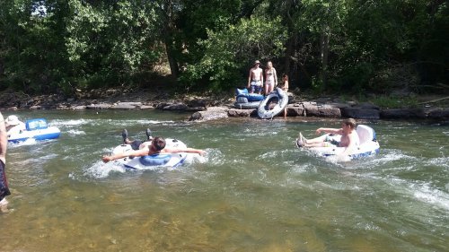 Two people float on inner tubes in a river, while others relax on the shore and nearby rocks. Lush greenery surrounds the scene.