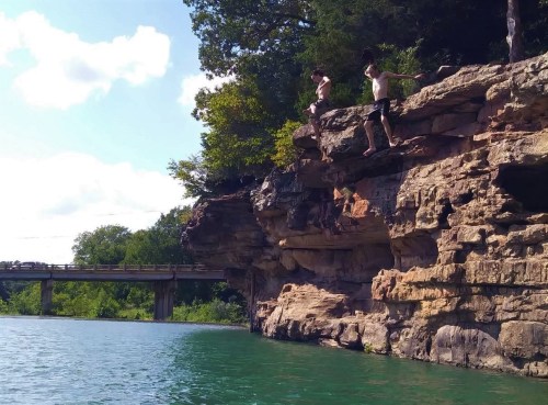 Two people stand on a rocky cliff above a clear blue lake, preparing to jump into the water below.