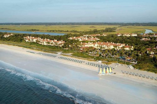 Aerial view of a beach with sailboats, lounge chairs, and a resort surrounded by greenery and water.