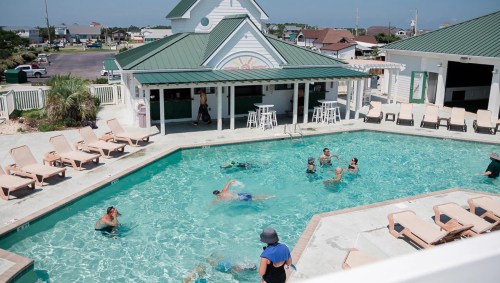 A sunny pool scene with people swimming and lounging around, featuring a bar area and beach chairs.