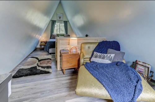 Cozy A-frame interior with two beds, a chair, and a window, featuring warm colors and a soft rug.
