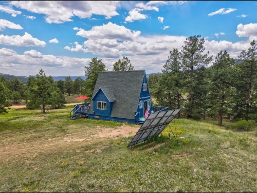 A blue house with a sloped roof surrounded by trees, featuring solar panels in the foreground and a cloudy sky above.