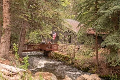 A cozy cabin by a flowing stream, surrounded by tall trees, with a deck and an American flag visible.
