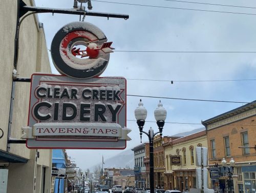 Sign for Clear Creek Cidery with a vintage design, hanging above a snowy street lined with historic buildings.