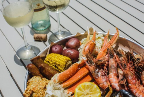 A seafood platter with shrimp, crab, corn, potatoes, and sausages, accompanied by two glasses of white wine.