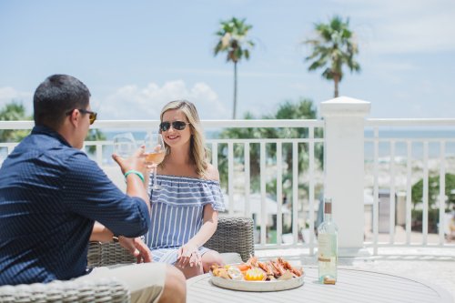 A couple enjoys drinks and snacks on a sunny patio with palm trees and a beach in the background.