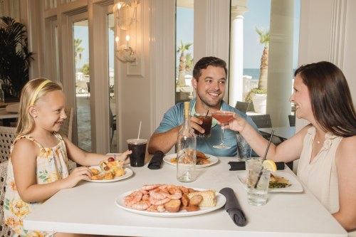 A family enjoys a meal at a seaside restaurant, toasting with drinks and sharing seafood dishes.