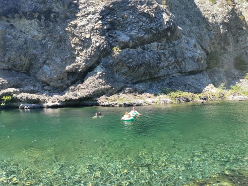 Two people swim in a clear, greenish-blue pool surrounded by rocky cliffs and lush greenery.