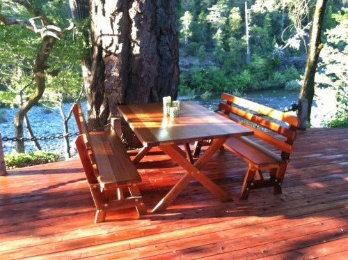 A wooden table and benches on a deck surrounded by trees, overlooking a river. Sunlight filters through the leaves.