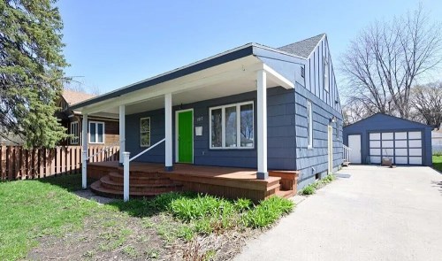 A blue house with a green door, front porch, and a garage, surrounded by grass and trees on a sunny day.