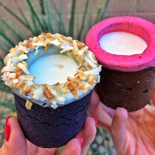 Two colorful dessert cups, one with a coconut topping and the other pink, held in hands against a green background.