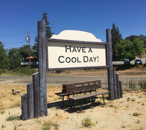 A wooden sign reads "Have a Cool Day!" with a bench in front, set against a clear blue sky and green trees.