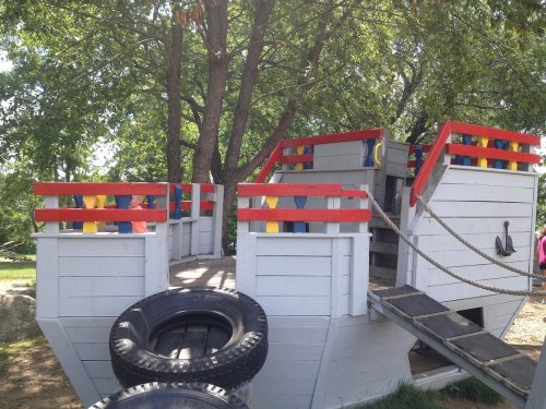 Colorful wooden play structure resembling a ship, with a tire and rope, set under a tree.