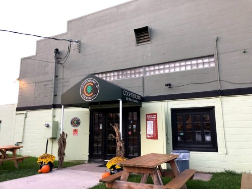 Exterior of Cooperstown Brewing Company, featuring a green building, entrance with signage, and outdoor seating.