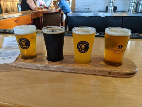 Four beer samples in glasses on a wooden flight board, with a brewery bar in the background.