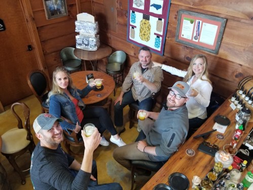 A group of five friends sitting at a wooden table, raising drinks and smiling in a cozy, rustic setting.
