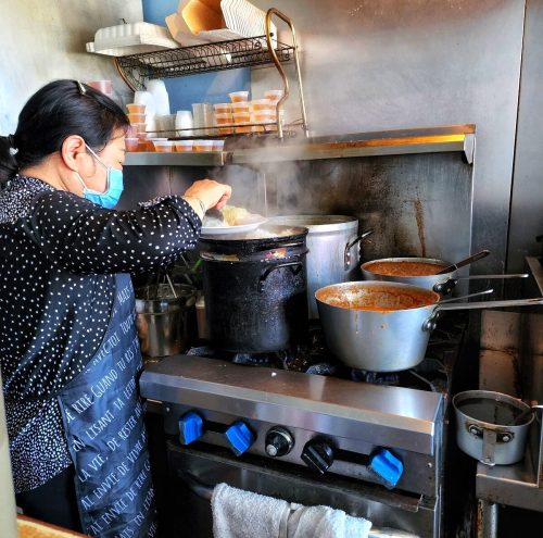 A woman in a mask stirs a pot on a stove in a busy kitchen, with steam rising and various pots on the burners.