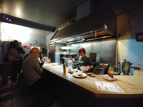 A cozy kitchen scene with a chef preparing food, a customer at the counter, and others in the background.