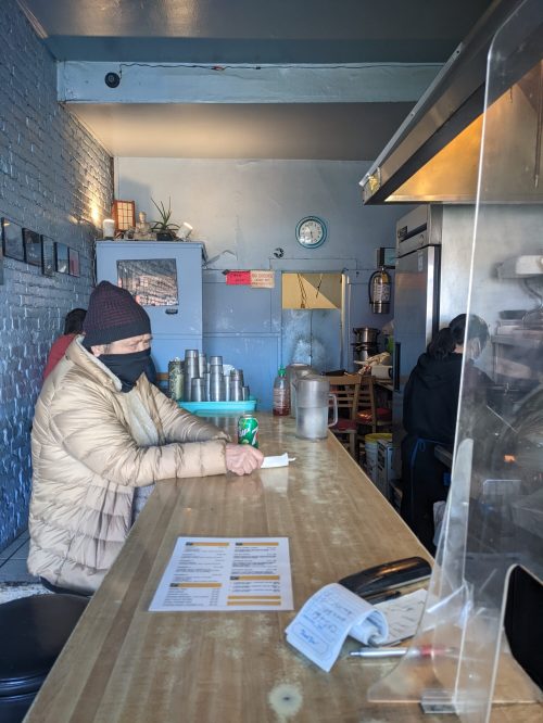 A person in a mask sits at a counter in a small restaurant, with a menu and kitchen visible in the background.