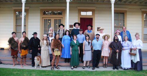 A large group of people in historical costumes poses in front of a vintage house, with a dog in the foreground.