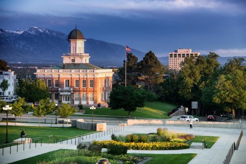 Historic building with a dome, surrounded by greenery and flowers, against a backdrop of mountains and a cloudy sky.