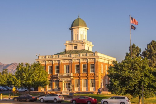 Historic building with a green dome, surrounded by trees and parked cars, under a clear sky with an American flag.