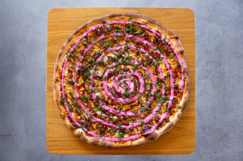 A large pizza topped with spiral patterns of pink sauce, herbs, and meat on a wooden table.