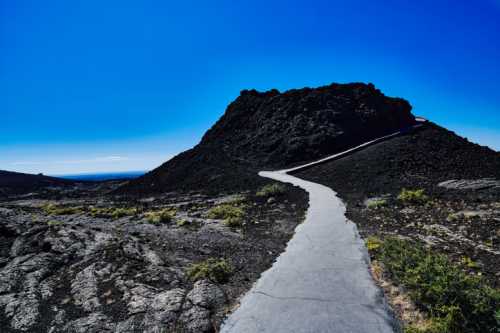 A winding path leads up a rocky hill against a clear blue sky, surrounded by volcanic terrain and sparse vegetation.