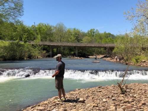 A man stands by a river with a small waterfall, while others fish and enjoy the sunny day in the background.