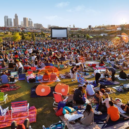 A large crowd gathers on a grassy field for an outdoor movie screening, with city skyline in the background.