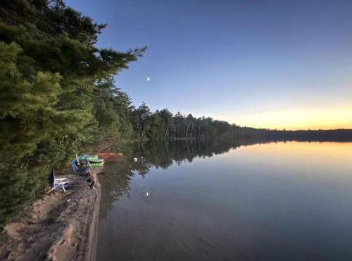 A serene lakeside view at dusk, with trees reflecting on calm water and a crescent moon in the sky.