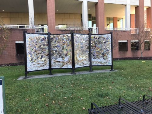 A large outdoor mural featuring a tree design with colorful leaves, set against a brick building and green grass.