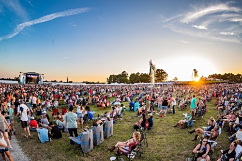 A large crowd enjoys a music festival at sunset, with a stage in the background and people seated on the grass.