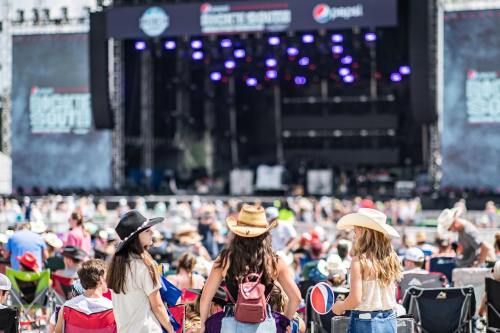 Three girls in cowboy hats enjoy a music festival, facing a large stage with colorful lights and a crowd in the background.