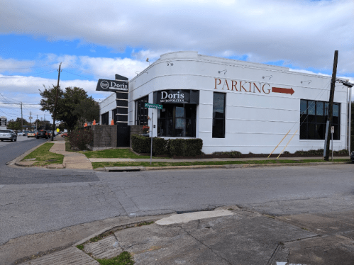 A white building with "Doris" signage and a parking arrow, located at a street corner with cars and trees nearby.