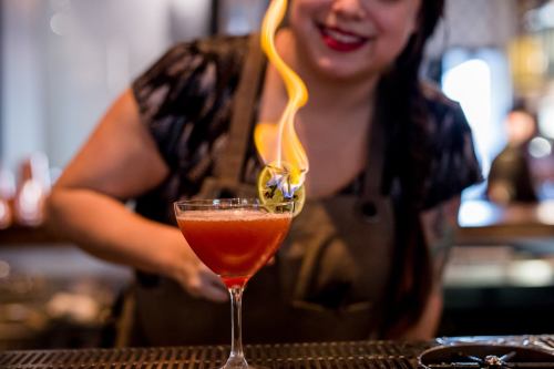 A bartender ignites a cocktail with a flame, showcasing a vibrant drink in a stylish glass.