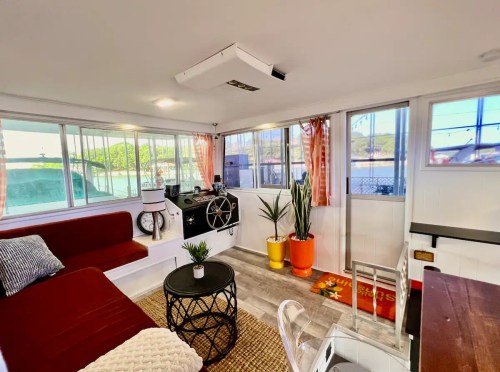 Cozy interior of a bright room with a red sofa, plants, and a view of water through large windows.