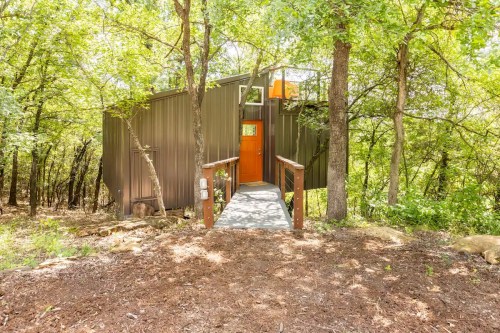 A modern cabin nestled among trees, featuring a wooden walkway and an orange door.