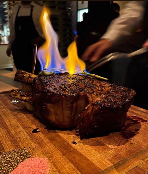 A large, grilled steak on a wooden board, with flames rising from the top, surrounded by seasoning.