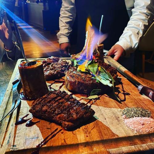 A wooden platter with various grilled meats, garnished with herbs, and a flame rising from one of the dishes.