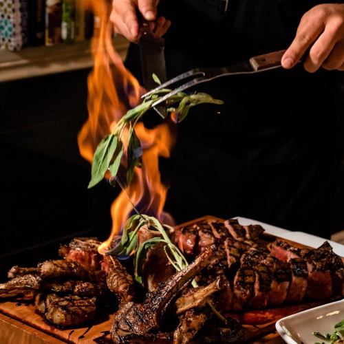 A chef adds herbs to a flaming grilled steak and shrimp on a wooden cutting board.