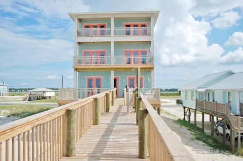 A three-story beach house with coral-colored accents, accessed by a wooden walkway, under a blue sky with clouds.