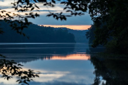 A serene lake at dawn, reflecting soft colors of the sky, framed by trees on the shore.