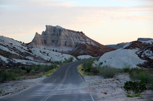 A winding road leads through a desert landscape with unique rock formations and soft, colorful hills under a pastel sky.