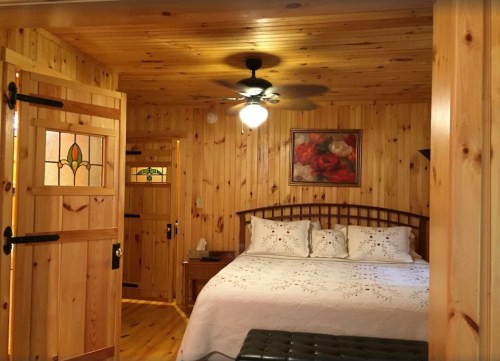 Cozy wooden bedroom with a queen bed, decorative pillows, and a ceiling fan, featuring warm wood paneling and art.