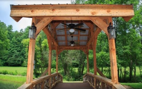 A wooden bridge with a covered structure, featuring ceiling fans and surrounded by lush greenery.