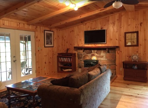 Cozy wooden living room with a stone fireplace, TV, and comfortable sofa, featuring large glass doors and warm lighting.